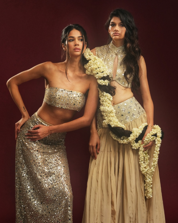 Two female models wearing contemporary festivewear from Alpha & Omega’s HER/HER collection, photographed against a burgundy studio backdrop.
